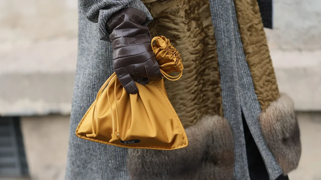 A mustard hued Prada Wish satin bag carried by a person wearing brown leather gloves, an oversized grey coat and brown fur scarf.