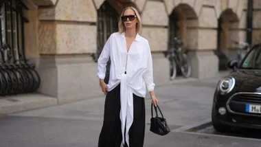 Woman wears white shirt and black elastic waist drawstring pants, sunglasses and a black top handle leather handbag.
