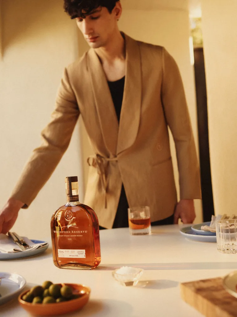 Man wears beige linen Host Tux jacket by COMMAS while laying a table with a bottle of Woodford Reserve whisky