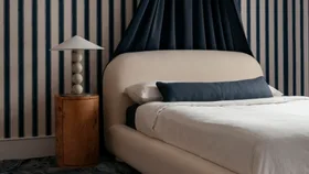 A cream-coloured upholstered bed with white bedding and navy pillows against a blue-and-white striped wall