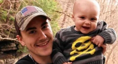 A man in a camouflage cap with an American flag patch holds a smiling baby in a dark sweater with a yellow skull design.