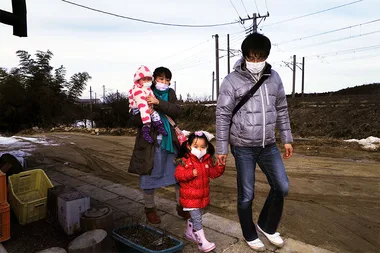 A family of four, wearing masks, walking outside in a rural area with dirt paths and power lines in the background.