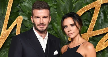 Two people pose together at an event, dressed formally, with a greenery and gold-textured background.
