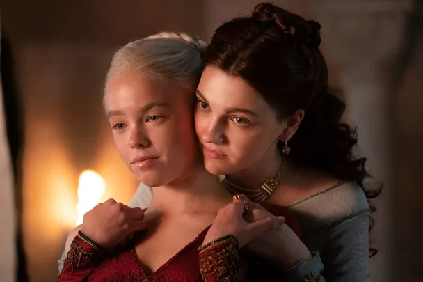 Two characters from "House of the Dragon" embrace tenderly, one with white hair and the other with dark brown hair.