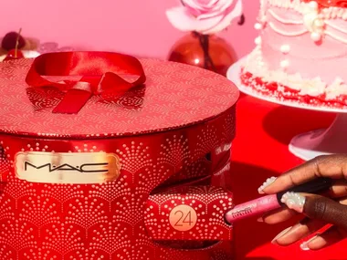 Hand holding MAC lipstick with red, festive MAC gift box and pink cake in the background.