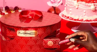 Hand holding MAC lipstick with red, festive MAC gift box and pink cake in the background.