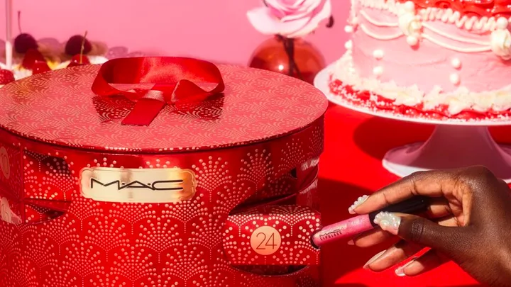 Hand holding MAC lipstick with red, festive MAC gift box and pink cake in the background.