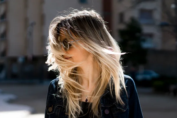 Woman with windblown hair covers her face outdoors, wearing sunglasses and a denim jacket.