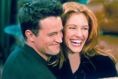 A man and woman laughing together closely, both wearing dark clothing, with blurred background.
