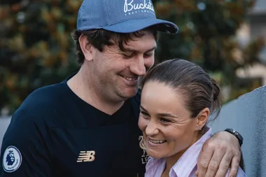 A man in a blue cap and sports shirt hugs a smiling woman in a striped shirt outdoors.