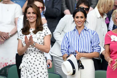 Two women smiling and clapping in a crowd; one in a white polka dot dress and the other in a blue striped shirt holding a hat.