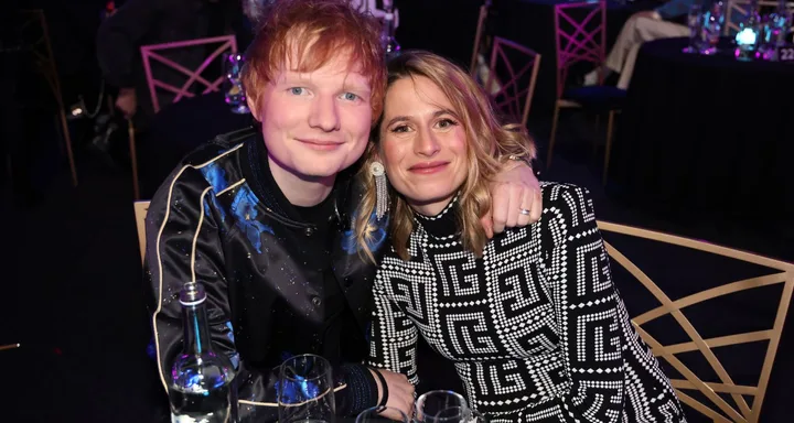 A man with red hair and a woman with blonde hair sitting closely together at a formal event, smiling at the camera.