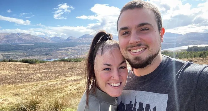 A smiling couple takes a selfie outdoors with mountains and a bright blue sky in the background.