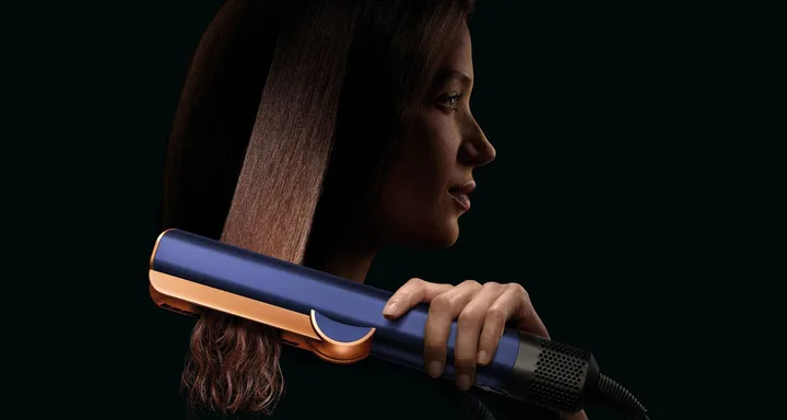 A woman using a blue and gold flat iron to straighten her hair against a dark background.