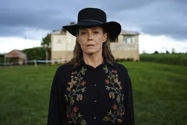 A woman in a black hat and floral embroidered shirt stands in a green field with a farmhouse in the background.