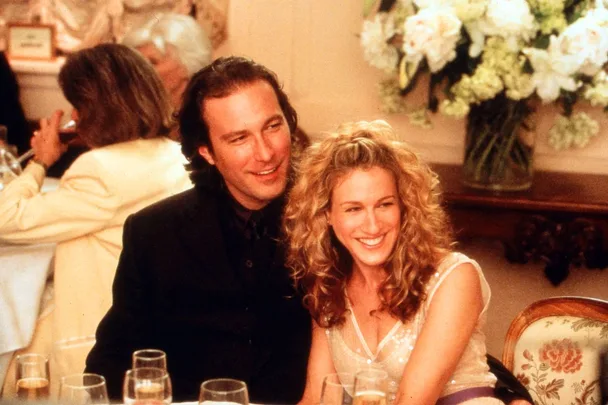 A man and a woman are sitting closely together at a formal event, smiling, with a flower arrangement behind them.