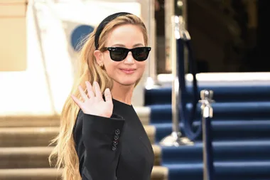 A woman wearing black sunglasses and a black headband waves and smiles while standing in front of a building.