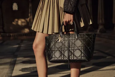 Person holding a black leather quilted handbag with gold monogram, wearing a beige pleated skirt and dark jacket.