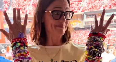 Woman smiles in crowded stadium, wearing numerous colorful beaded bracelets on both arms, hands held up.