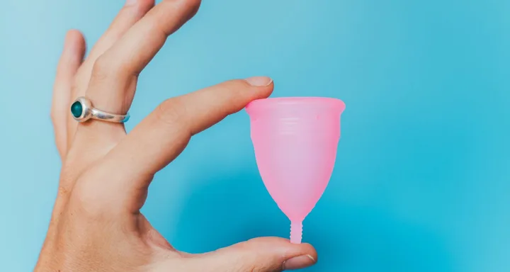 A hand holding a pink menstrual cup against a light blue background.