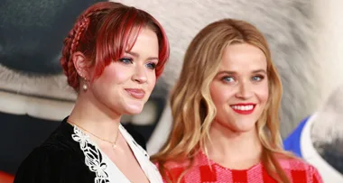 Two women smiling at a red carpet event, one with red hair and the other with blonde hair.