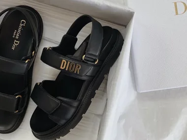 Black Dior sandals with gold-tone "DIOR" logo, black insoles with "Christian Dior Paris" text, in a white Dior box.