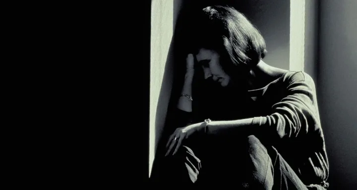 A woman sits in a dark corner, resting her head on her hand, seemingly overwhelmed and distressed.