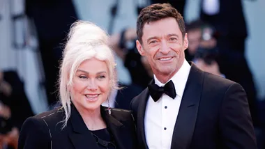 A woman with blonde hair and a man in a black tuxedo smile on the red carpet at an event.