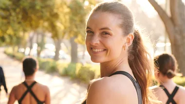 Woman smiling outdoors during a workout with a blurred background of sunshine and trees.