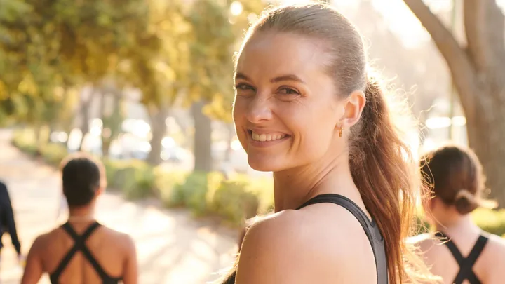 Woman smiling outdoors during a workout with a blurred background of sunshine and trees.