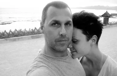 A couple taking a close-up selfie by the beach during sunset, with the ocean and mountains in the background.