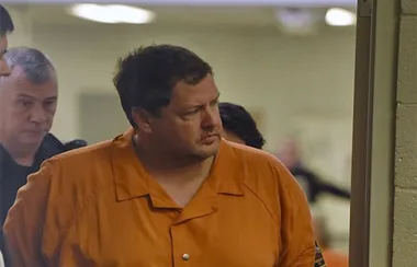 Man in orange prison uniform escorted by guards in a hallway.