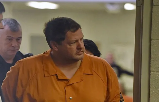 Man in orange prison uniform escorted by guards in a hallway.