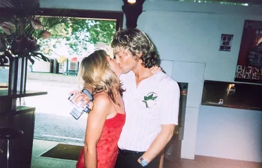 A couple kisses at an indoor location, with the man holding a camera and wearing a white shirt and the woman in a red dress.