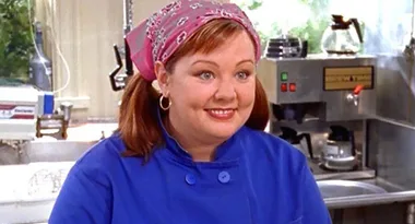 A woman in a blue chef's jacket and pink bandana smiles in a kitchen setting with coffee machines in the background.