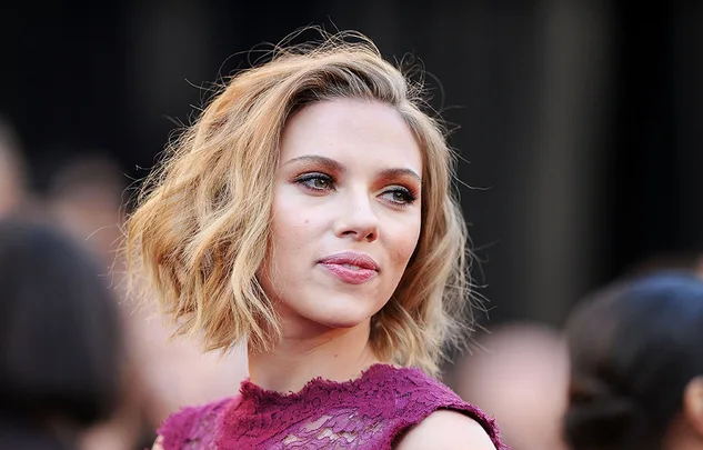 A person with short wavy blond hair wearing a lacy, sleeveless red top, looking to the side in a formal setting.