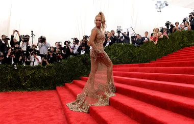 A person poses on red carpet stairs in a transparent, embellished gown as photographers take pictures.