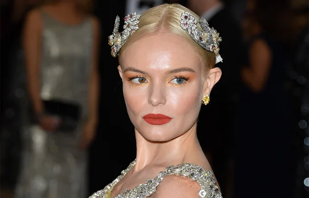 A woman with slicked-back blonde hair adorned with jeweled hairpieces, wearing bold red lipstick and gold eyeshadow at a formal event.