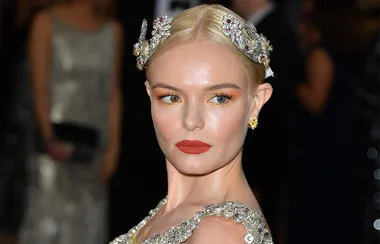 A woman with slicked-back blonde hair adorned with jeweled hairpieces, wearing bold red lipstick and gold eyeshadow at a formal event.