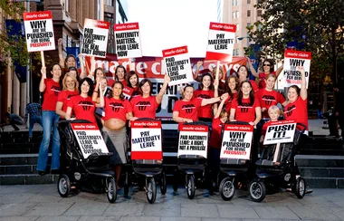 Group of women in red shirts with strollers holding signs advocating for paid maternity leave on city steps.
