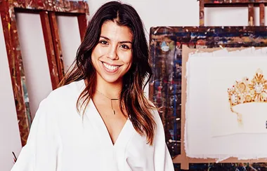 Woman in a white shirt smiling in an art studio with paintings in the background.