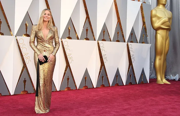 A woman in a gold dress on the Oscars red carpet, with an Oscar statue in the background.