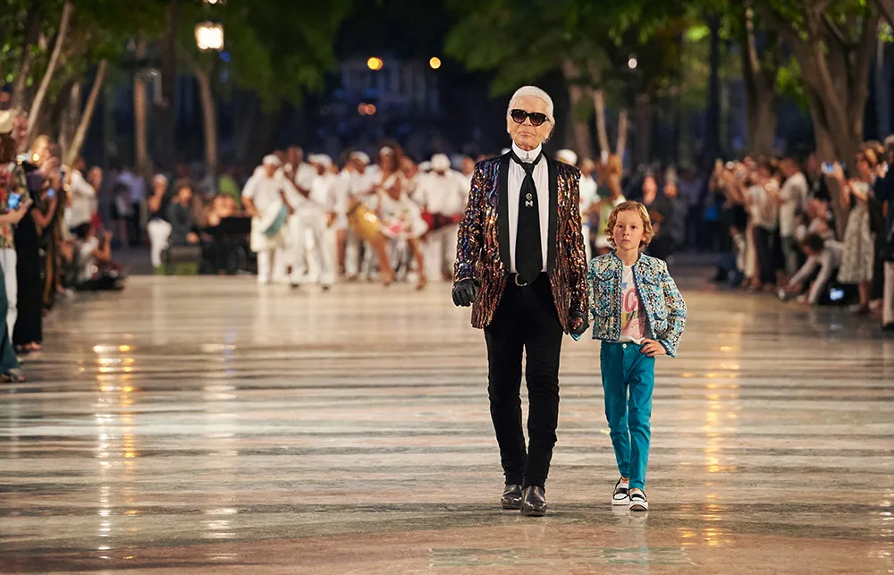 Fashionable man and child walking confidently in a lively outdoor setting, with a crowd in the background.