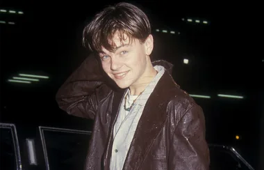 Young person in a leather jacket smiling, with tousled hair, posing outdoors at night near a vehicle.