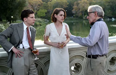 Three individuals are engaged in conversation on a bridge overlooking a lake, with drinks in hand, likely on a film set.