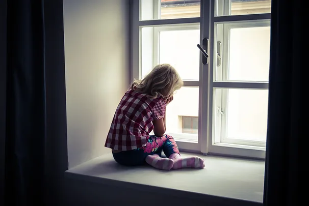 Young child in checkered shirt and colorful leggings sitting on a windowsill, facing away and looking out the window.