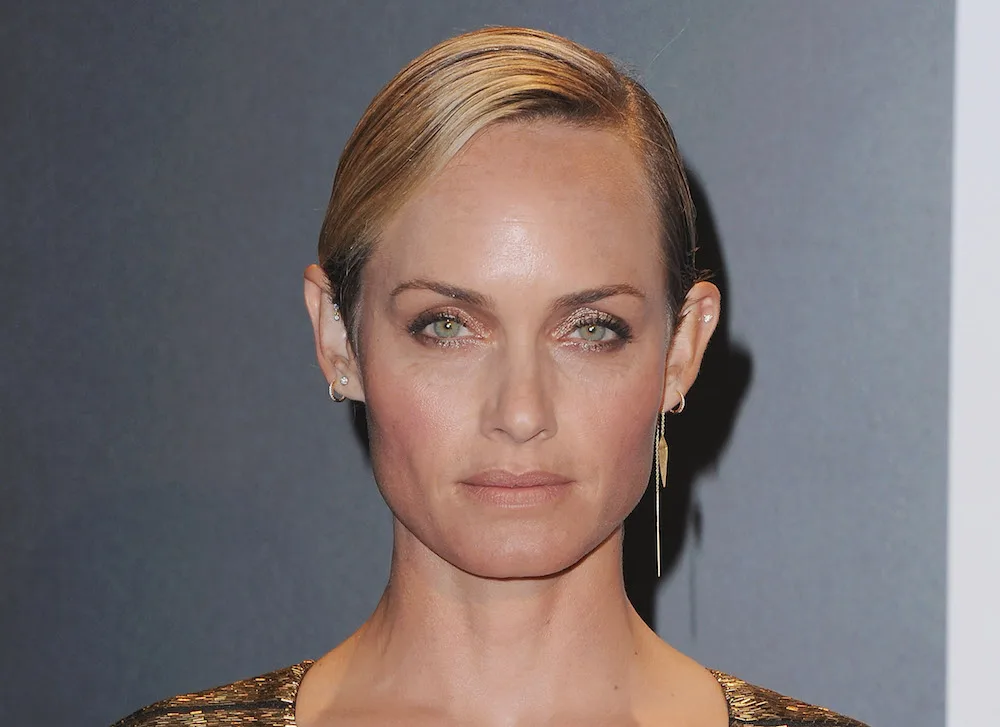 A person with short, light brown hair and wearing a gold dress, standing against a neutral background.