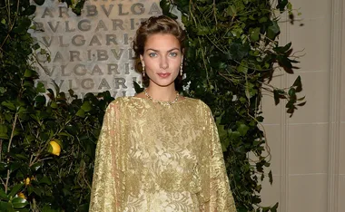 Woman in gold lace dress poses at a BVLGARI event, surrounded by green foliage and a branded backdrop.