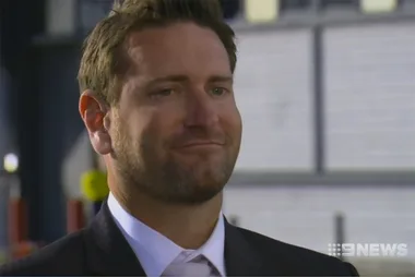 A man in a suit with a faint smile, standing outdoors. The image is credited to 9 News.