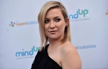 A woman with short blonde hair poses in front of a branded backdrop, wearing a black one-shoulder dress.
